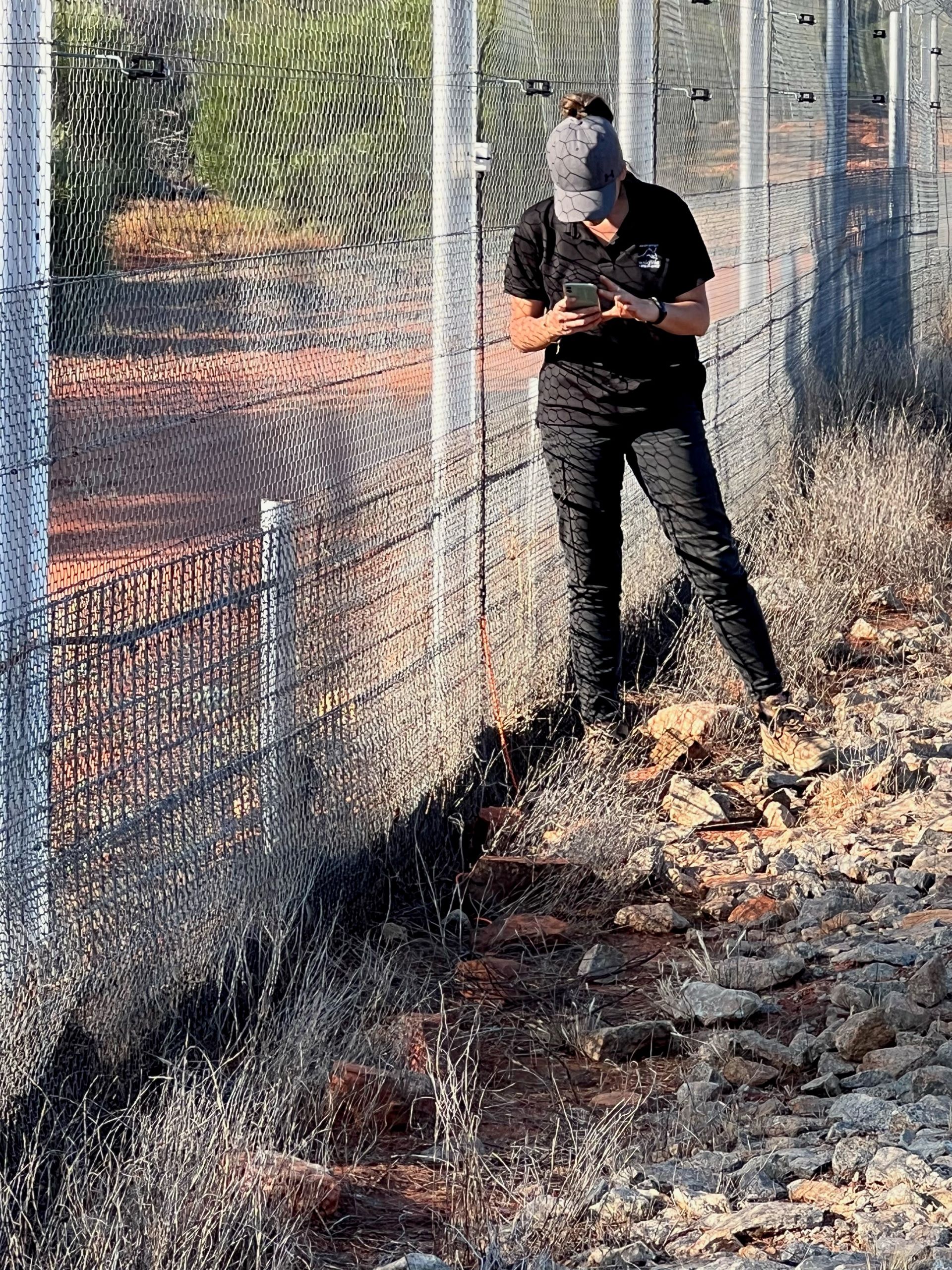 A young woman stands in front of a grey fence looking at a handheld device